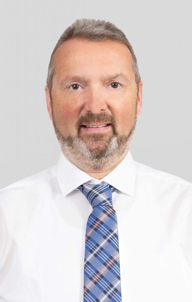 Ein Portrait eines Mitarbeiters mit gräulichen Haaren und Bart, weißem Hemd und blau-karierter Krawatte