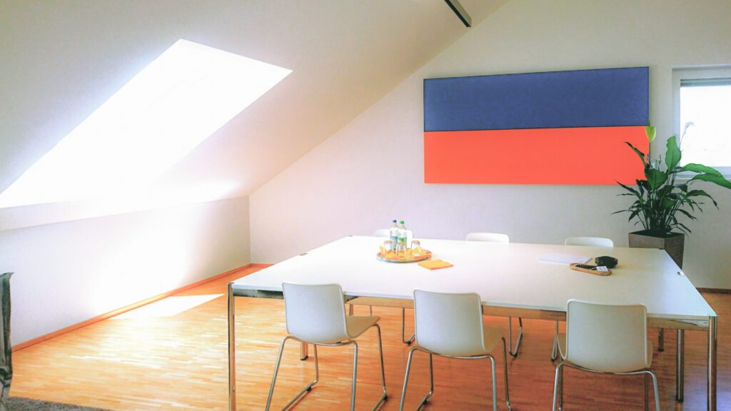 Ein Konferenzraum mit einem großen weißen Tisch mit Stühlen drumherum, durch ein Dachfester scheint die Sonne. An der Wand ist ein Fernseher beschäftigt mit einm blauen und einem roten Streifen als Hintergrund.